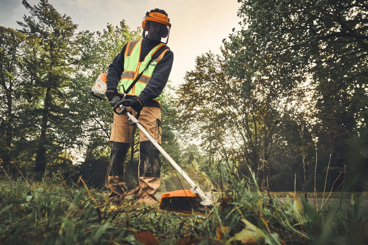 A groundsman in hi-vis cutting long grass in a park with a STIHL KMA 200 R and brushcutter attachment